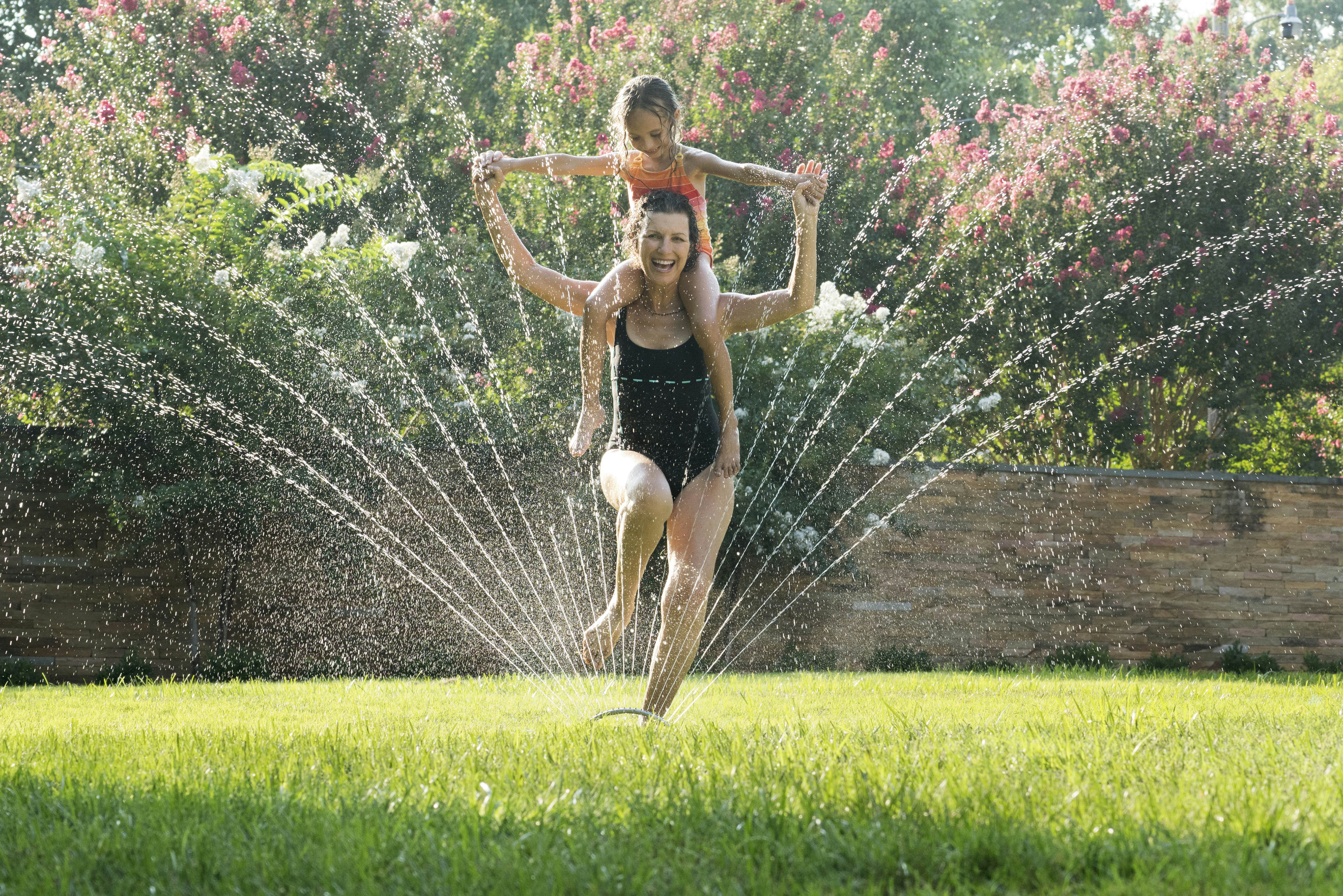 Mixed Race grandmother carrying granddaughter on shoulders in sprinkler